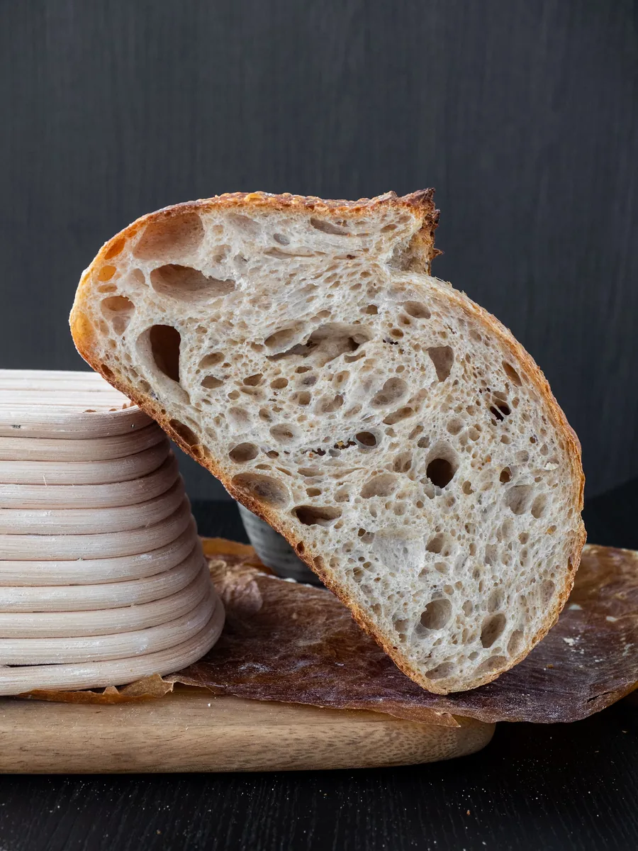 A slice of sourdough with an open, airy crumb, leaning against a stack of proofing baskets